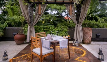 Un área de comedor al aire libre con una mesa dispuesta para dos personas, rodeada de vegetación y una pérgola.