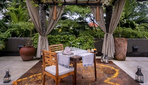 An outdoor dining area with a table set for two, surrounded by greenery and a pergola.