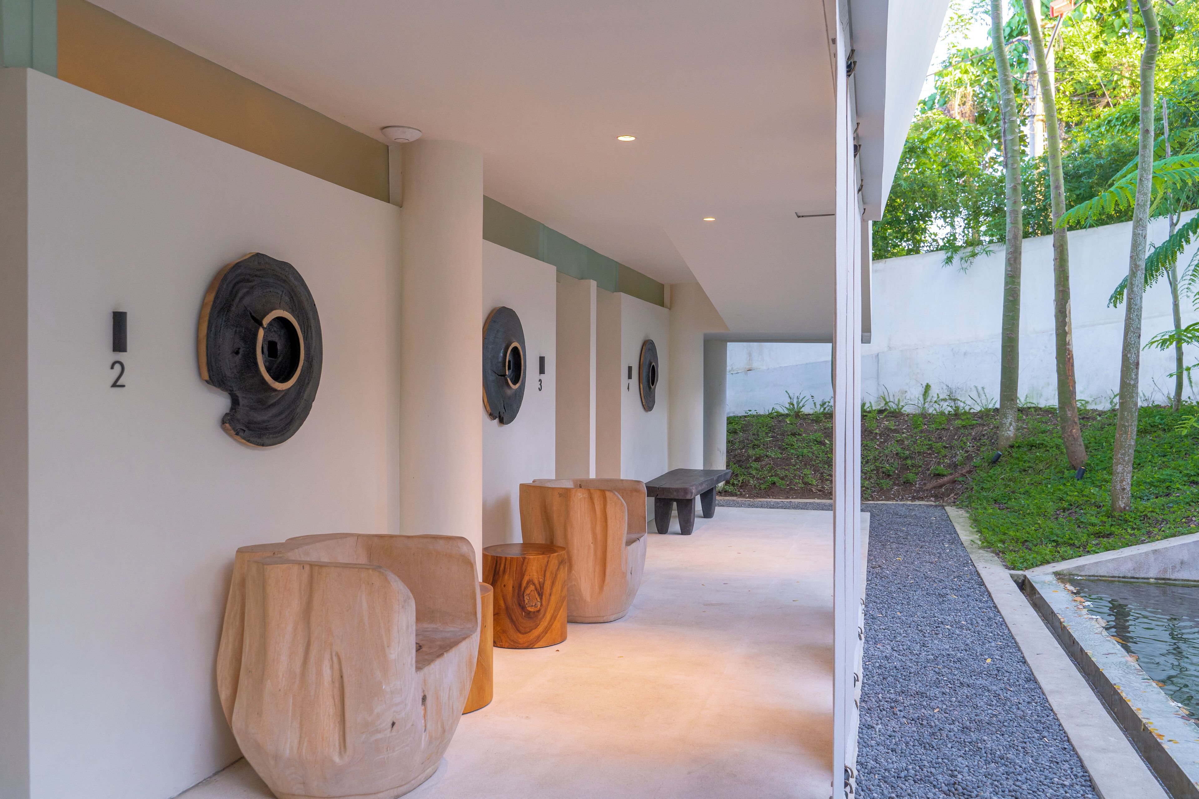 A corridor with wooden furniture and wall-mounted circular designs, leading to an outdoor area with a pool and greenery.