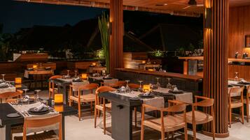 An outdoor dining area with multiple tables and chairs, wooden columns, and a bar area.