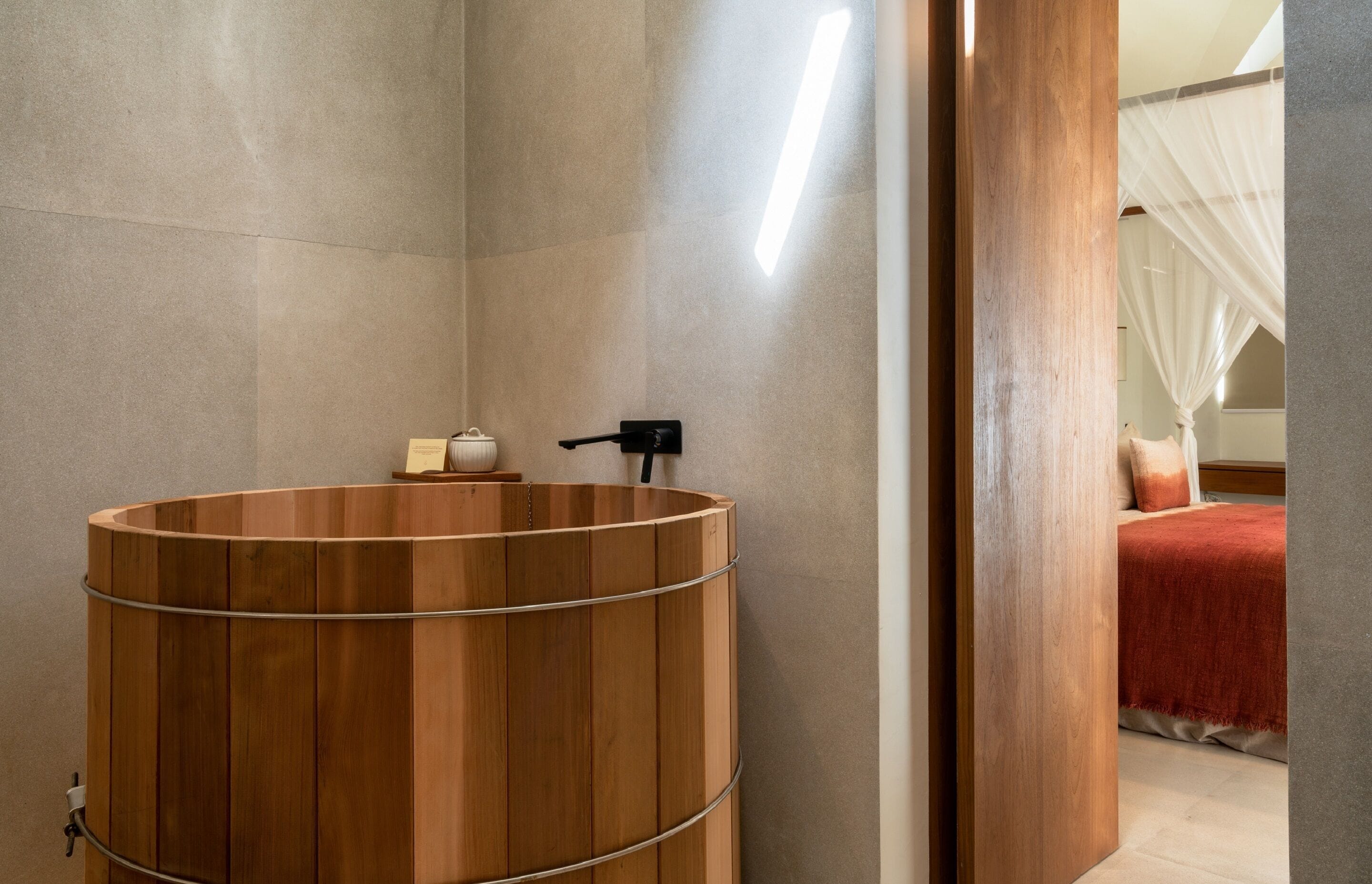 A modern bathroom with a wooden bathtub, a towel rack, and a bed with a canopy in the background.