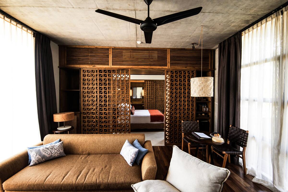 A hotel room with a sofa, a dining table, a ceiling fan, and a bed with a wooden headboard.