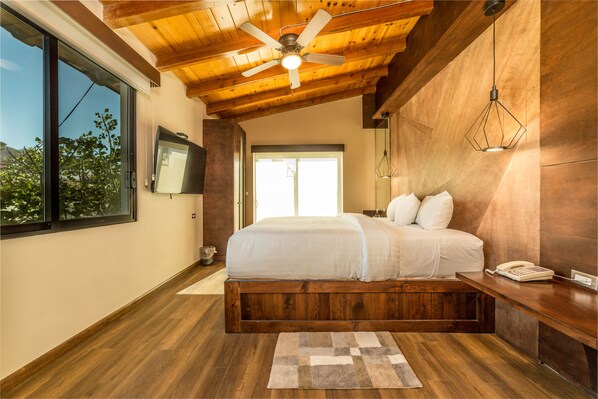 A bedroom with a wooden ceiling, a bed, a TV on the wall, and a window with a view. - Bottega Hotel Boutique (Valle de Guadalupe)