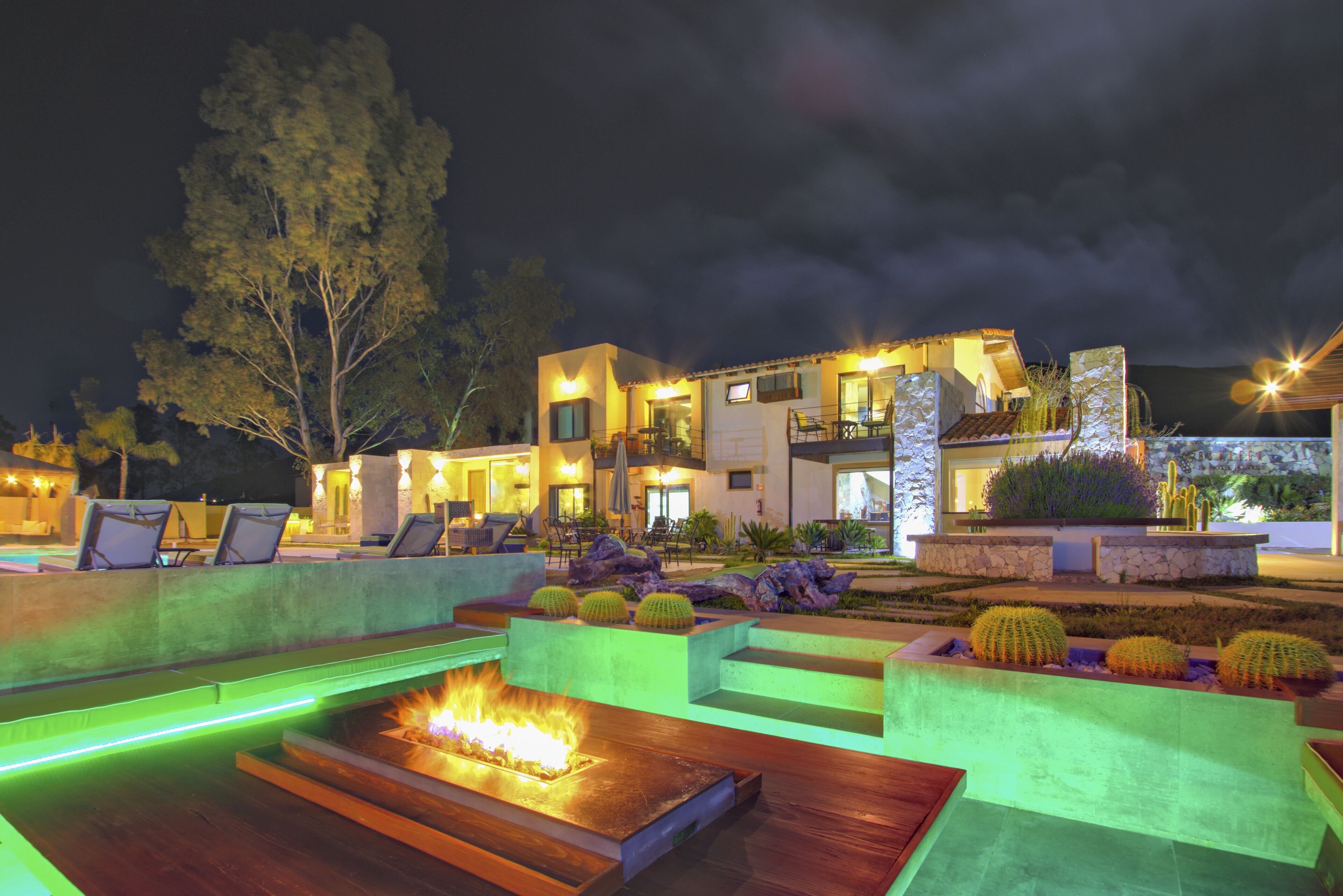 A modern outdoor seating area with a fire feature, surrounded by cacti and plants, and a building with a stone facade in the background.
