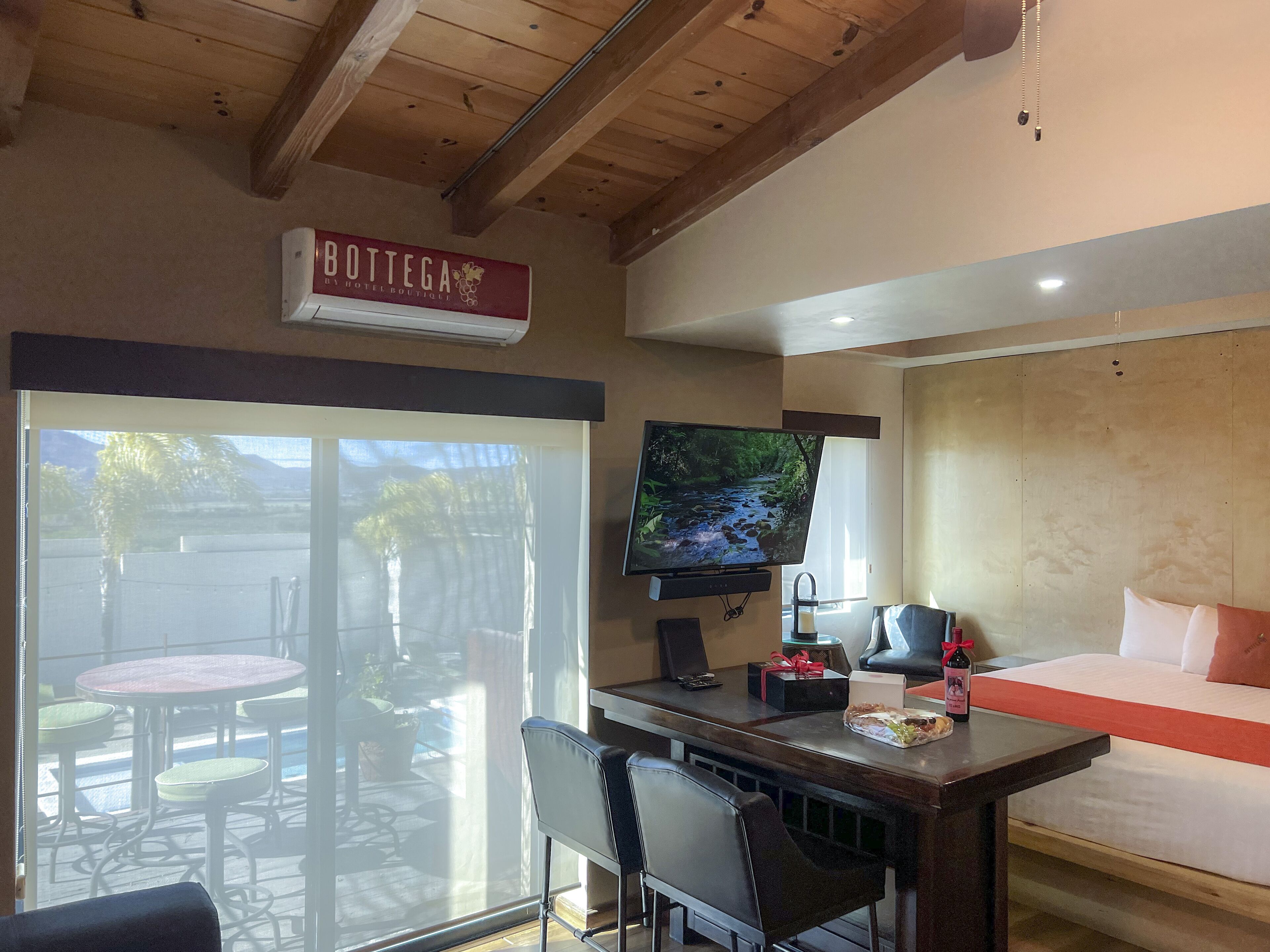 A room with a wooden ceiling, a television mounted on the wall, a bed with a red blanket, and a table with chairs.