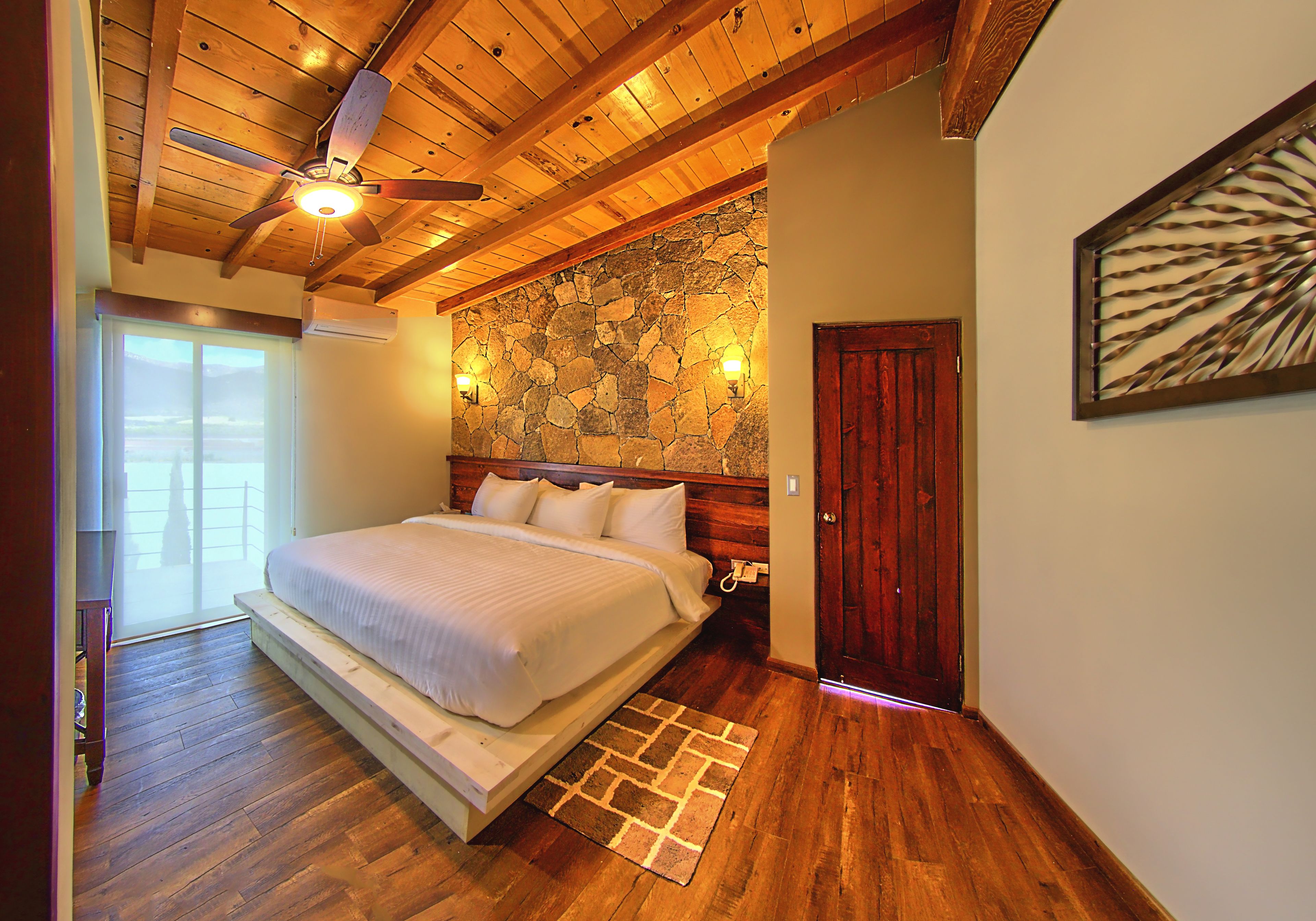 A bedroom with a wooden ceiling, a stone wall, a bed with white bedding, a wooden door, and a large window.