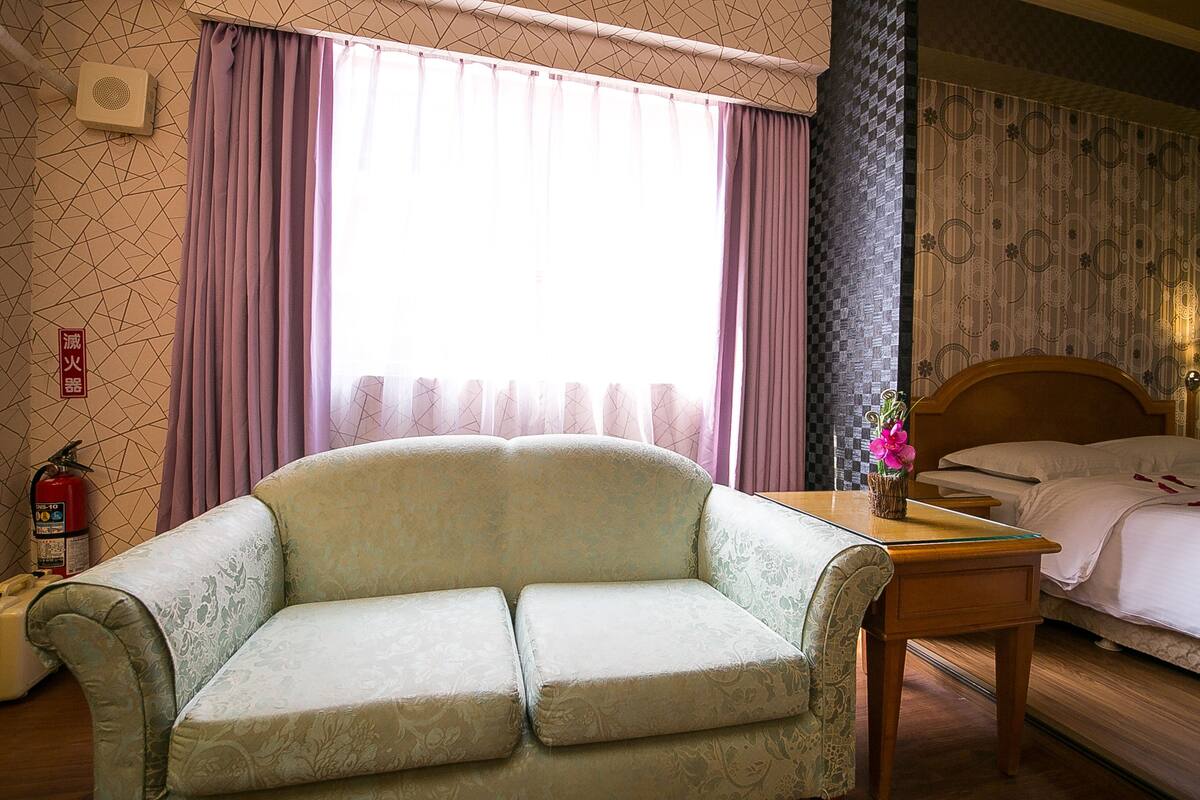 Et hotelværelse med sofa, et træbord med blomster i en vase, seng med hvide sengetøj og et vindue med gardiner.