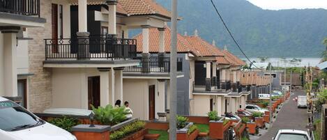 A residential street with houses featuring balconies and tiled roofs, a parked car, and a mountain backdrop.