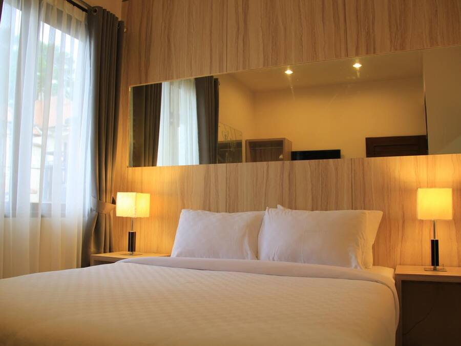 A hotel room with a large bed, two bedside tables with lamps, a large mirror above the bed, and sheer curtains on the window.
