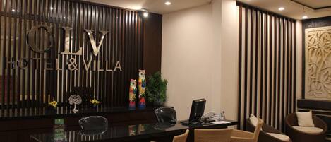 Uma receção com um letreiro a indicar "CLV HOTEL & VILLA". Há cadeiras de vime em frente à receção, uma planta em vaso e um painel decorativo na parede.