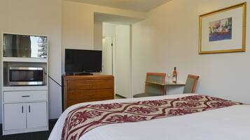 A hotel room with a bed, a television, a microwave, a small table with a bottle and glasses, and a framed picture on the wall.