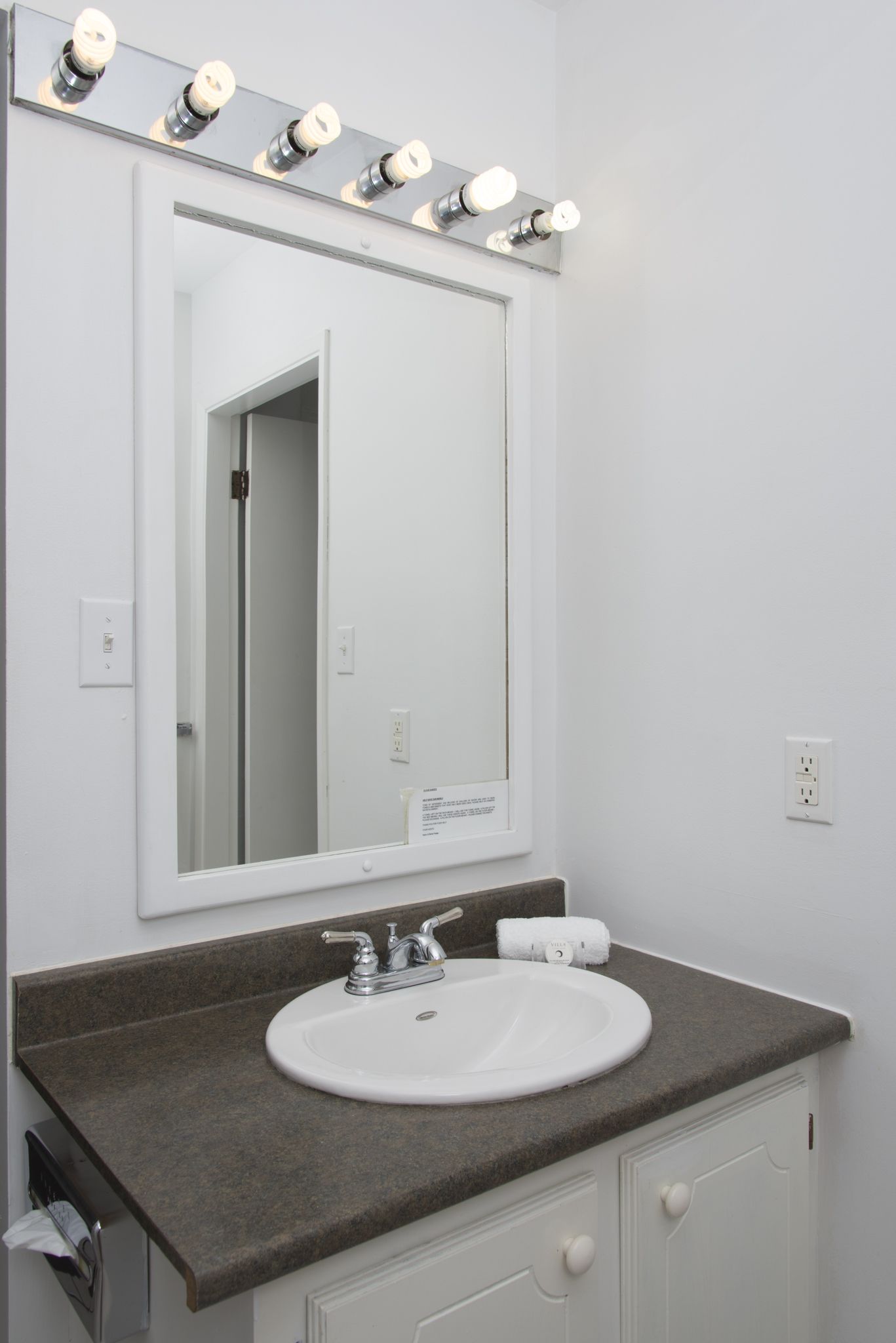 A bathroom with a white sink, a mirror with lights, and a towel dispenser.