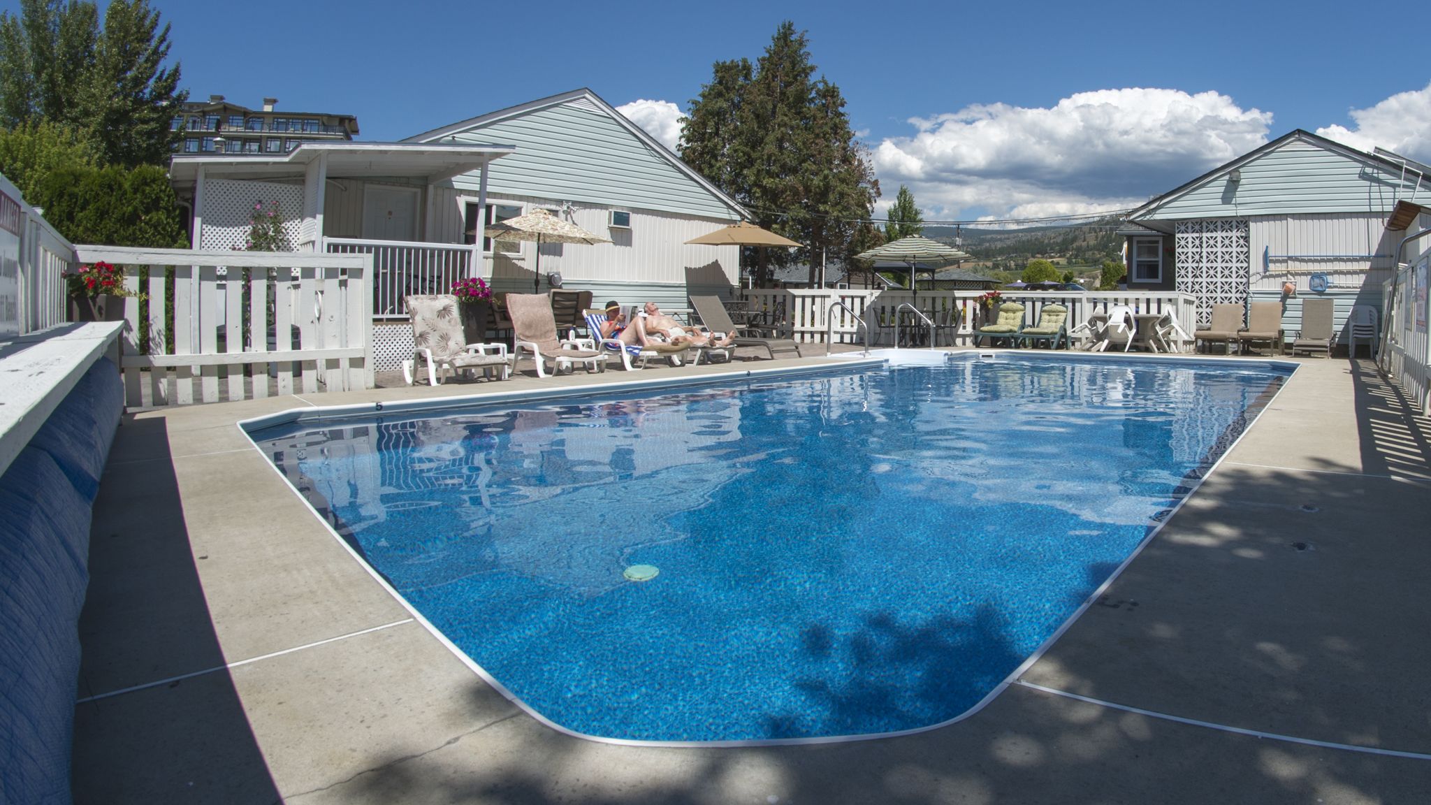 Una piscina rectangular con un deck blanco alrededor, un área de descanso techada con sillas y sombrillas, y una vista de casas al fondo.