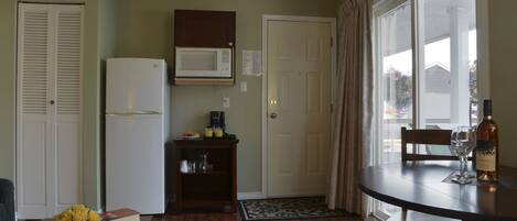Cocina compacta equipada con refrigerador, microondas y un pequeño rincón para café. Se aprecia una mesa de comedor con una botella de vino y una copa, junto a una silla. Una puerta da a un espacio exterior con vistas.