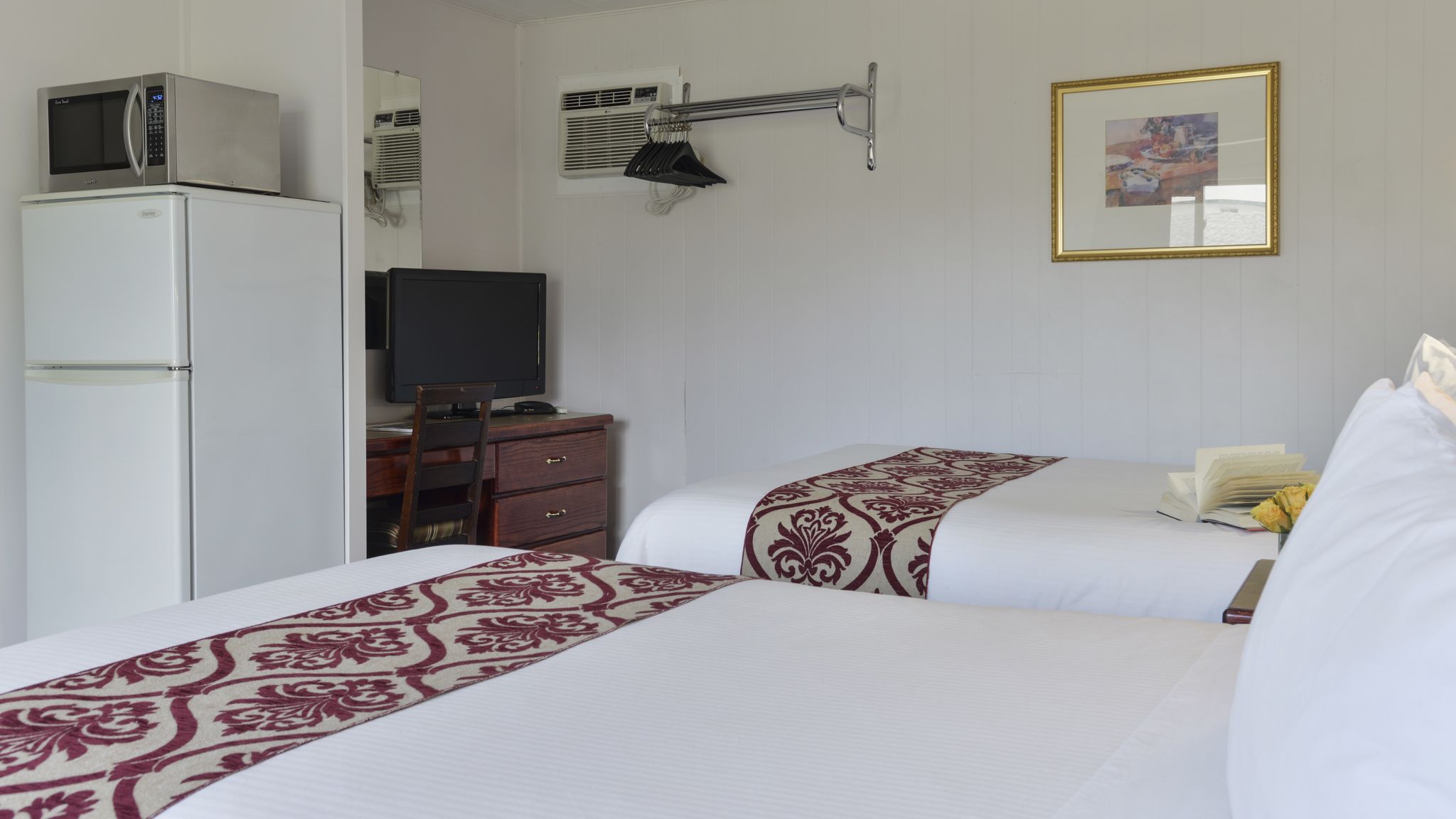 A hotel room with two beds, a refrigerator, a television, an air conditioning unit, and a framed picture on the wall.