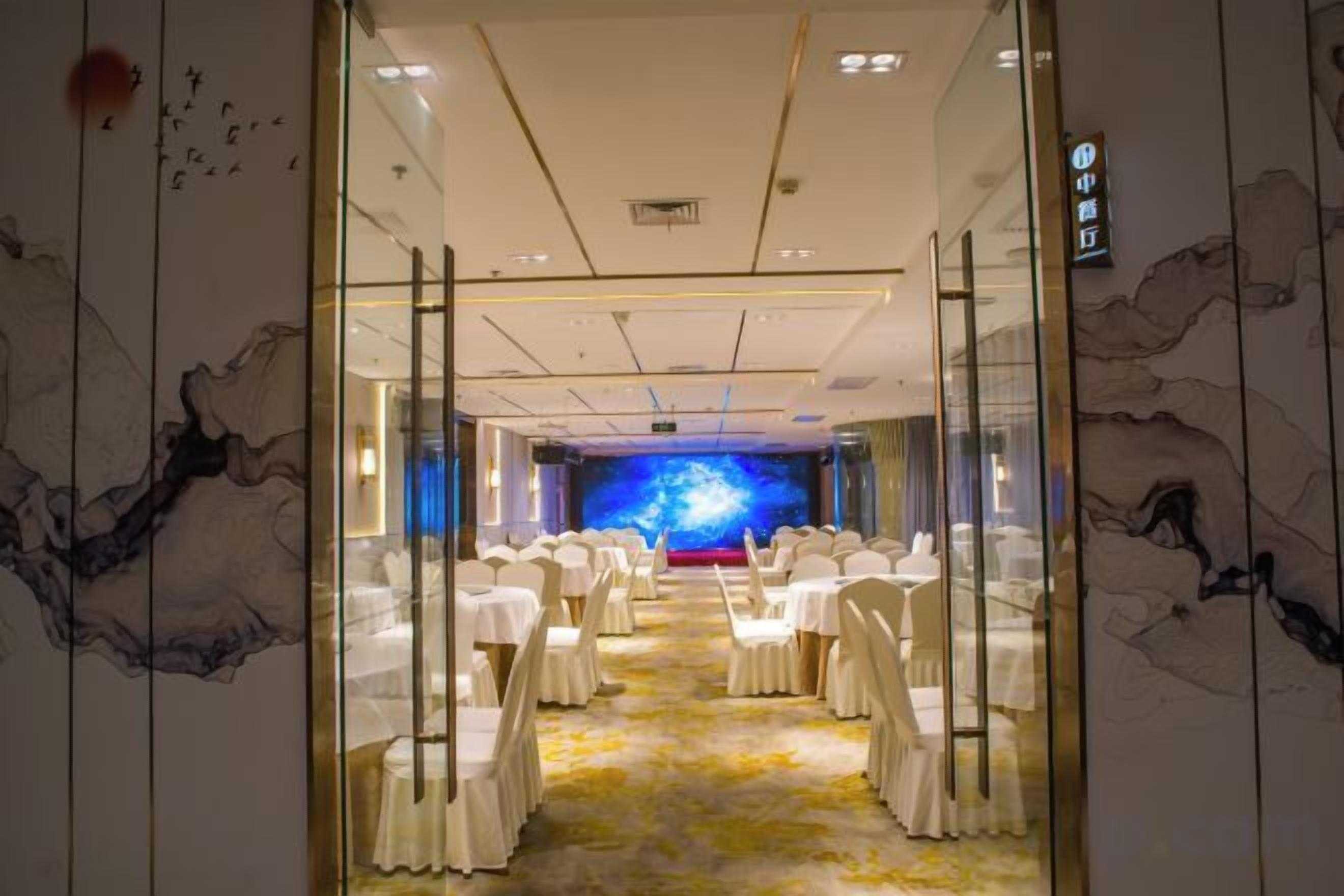 Sala de conferências de hotel com cadeiras brancas dispostas em fileiras, palco frontal e um grande mural na parede.