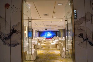 Un salón de conferencias de hotel con sillas blancas dispuestas en filas, un escenario al frente y un mural grande en la pared.
