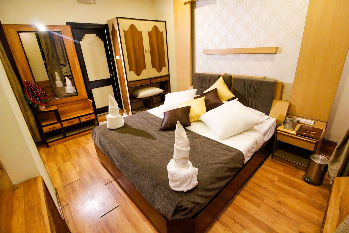 A hotel room with a large bed, a wooden wardrobe, a bedside table with a lamp, and a trash bin.