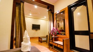 A hotel room with a wooden floor, a bed with a gray blanket, a wooden bench, a small table, a TV mounted on the wall, and a mirror with a wooden frame. - Hotel Rishi Regency (Jabalpur)