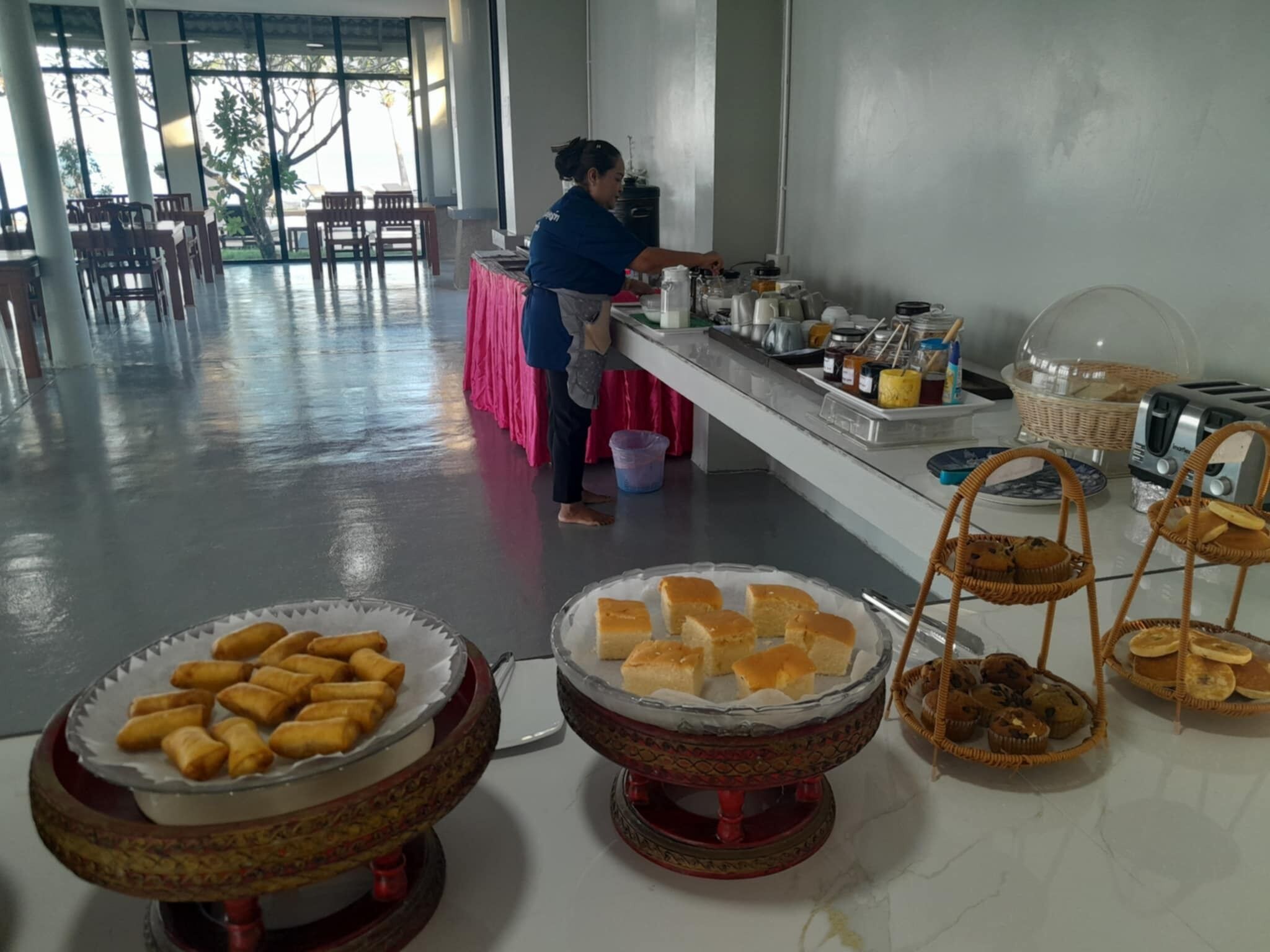 A buffet setup with various food items, a person serving, and a spacious room with tables and chairs.
