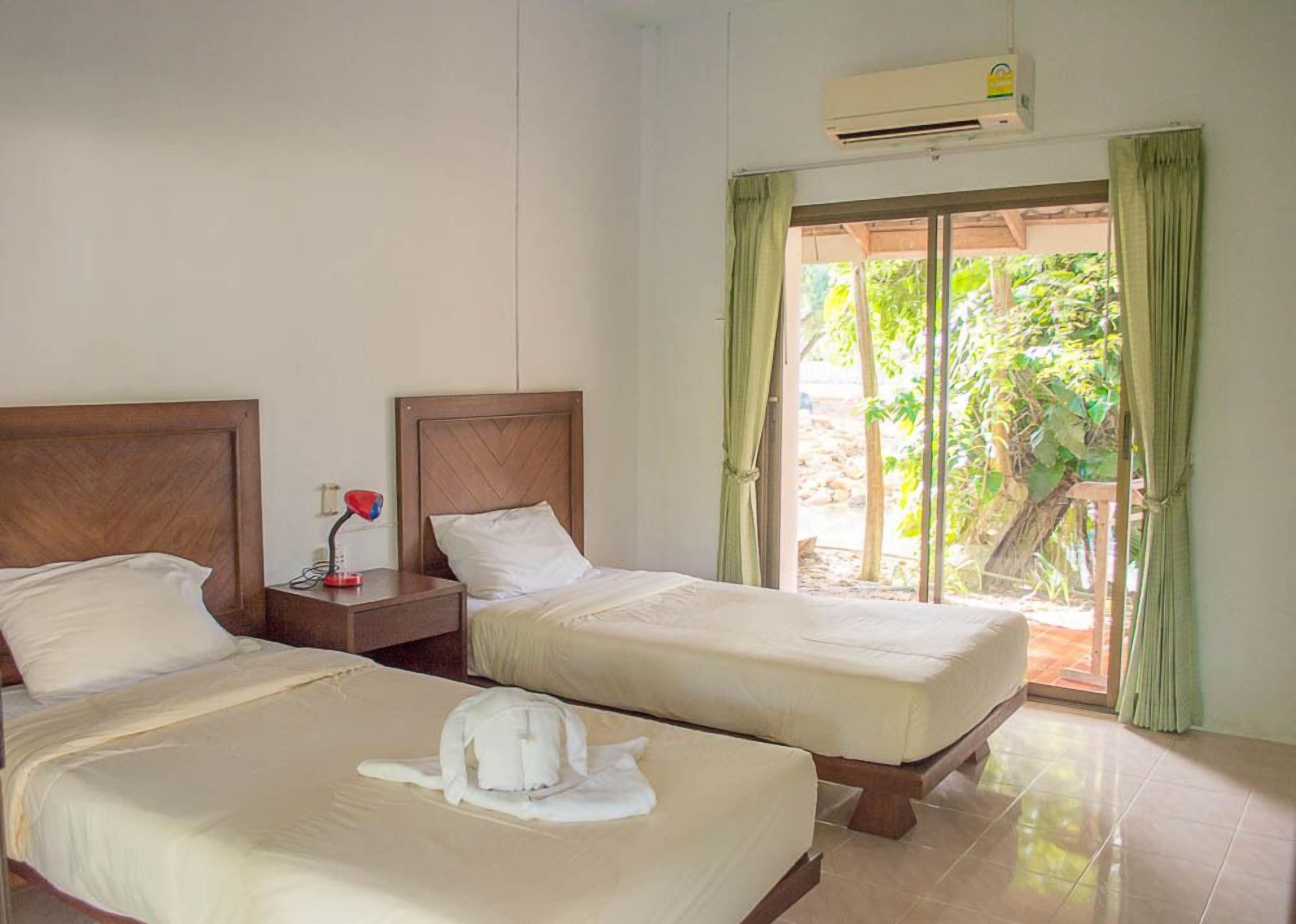 A hotel room with two beds, a nightstand, an air conditioner, and a view of greenery through a window.