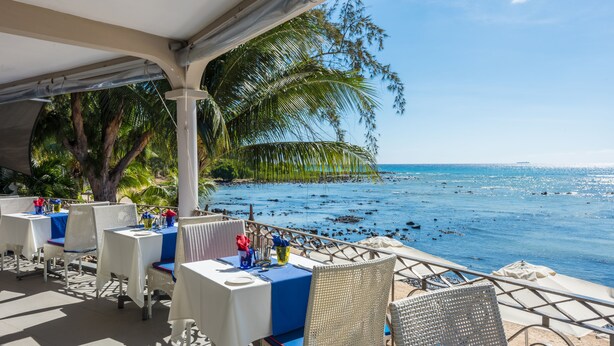 Un área de comedor al aire libre con mesas y sillas, con vista a una playa con palmeras y el océano.