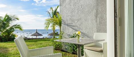 A balcony with wicker chairs and a table, overlooking a beach with palm trees and a thatched umbrella.