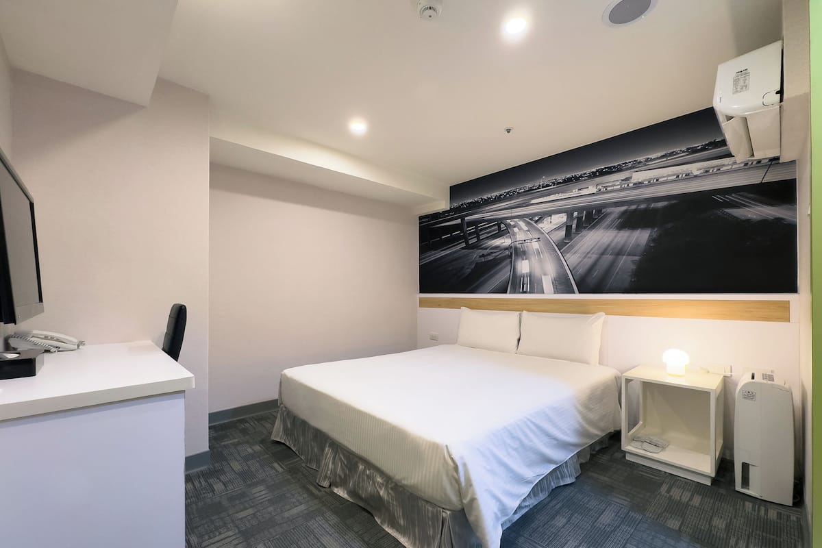 Una habitación de hotel con cama, escritorio con teléfono, mesita de noche con lámpara y una gran foto blanco y negro en la pared.