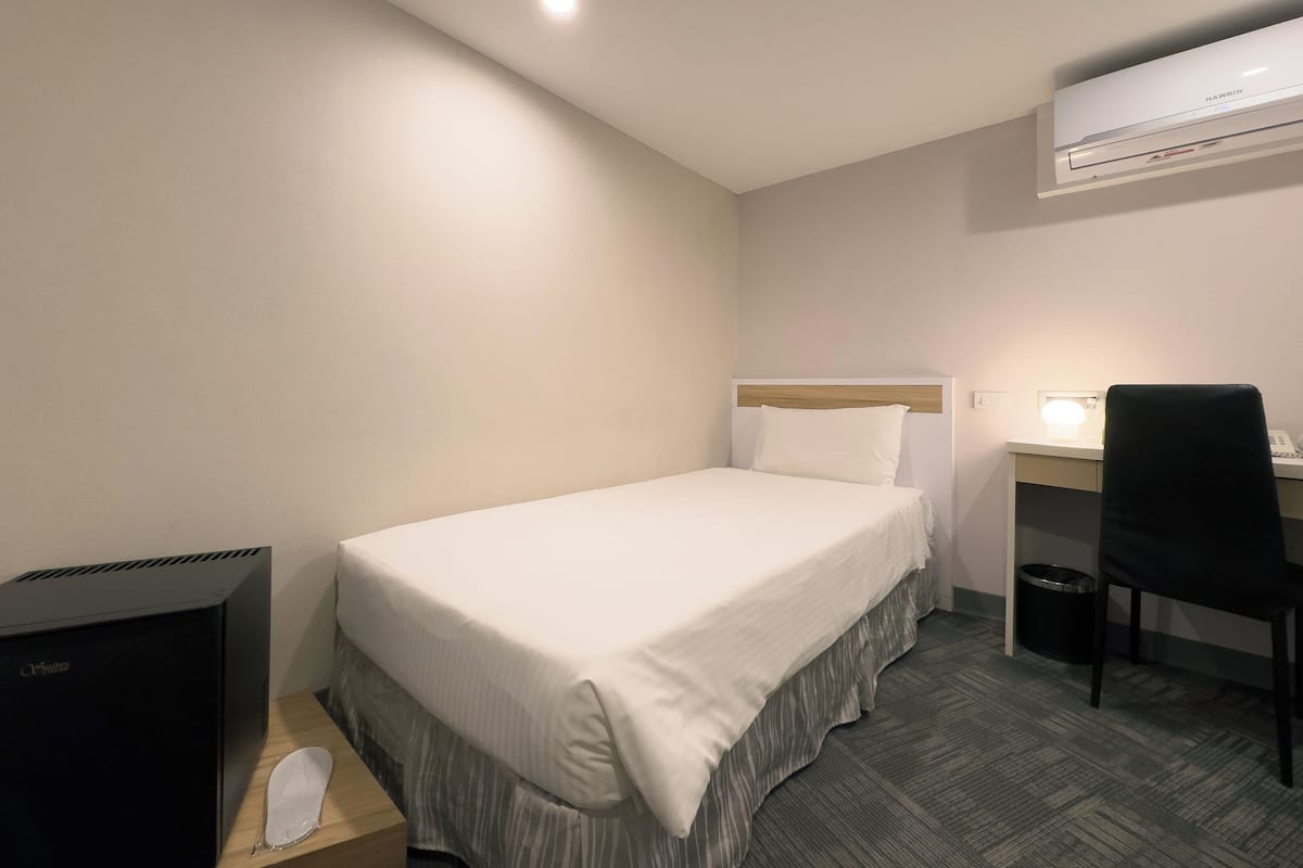 Una habitación de hotel con una cama individual, un escritorio con silla, un cubo de basura y un calentador negro.