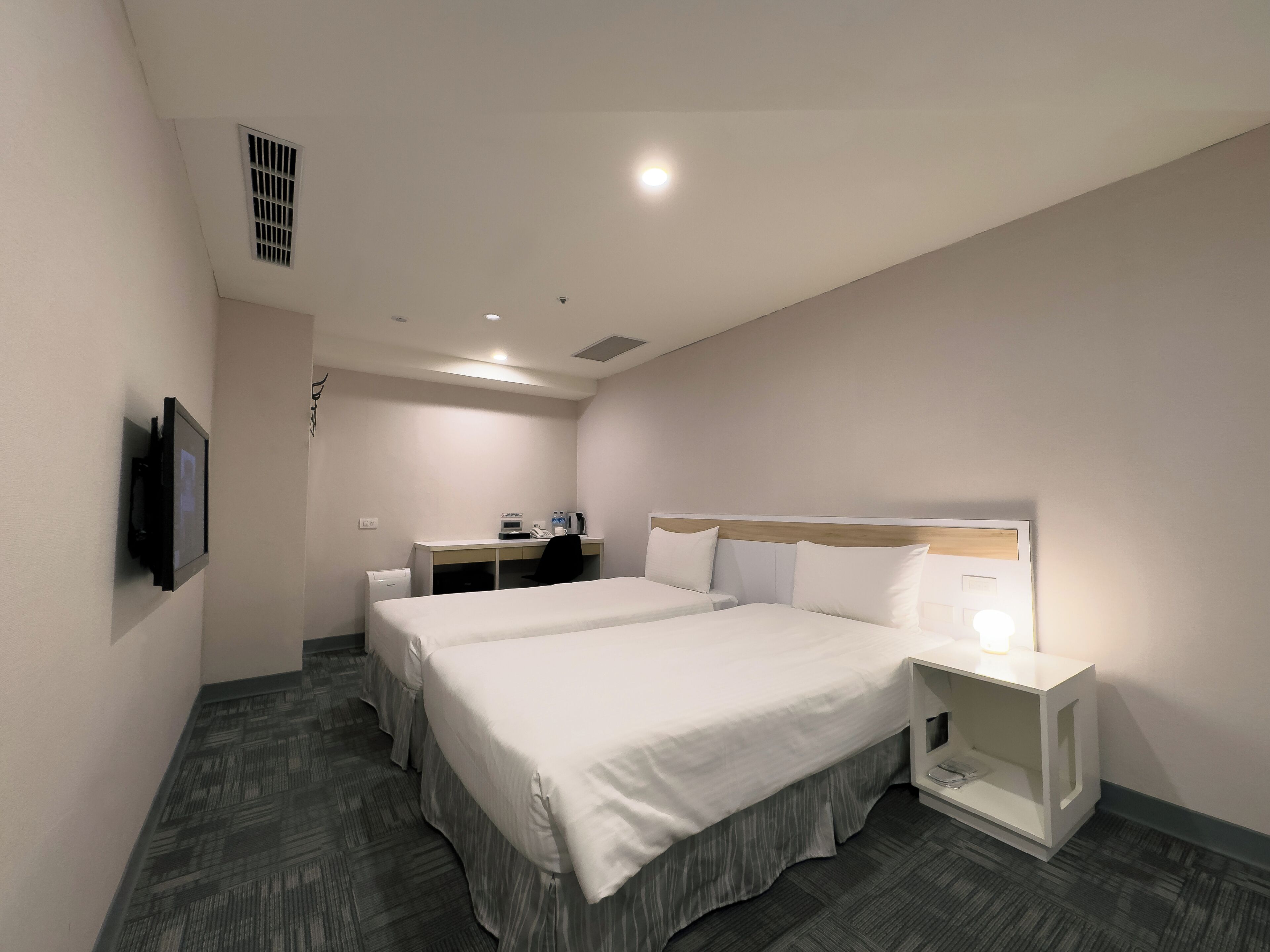 A hotel room with two beds, a desk, a chair, a TV, and a bedside table with a lamp.