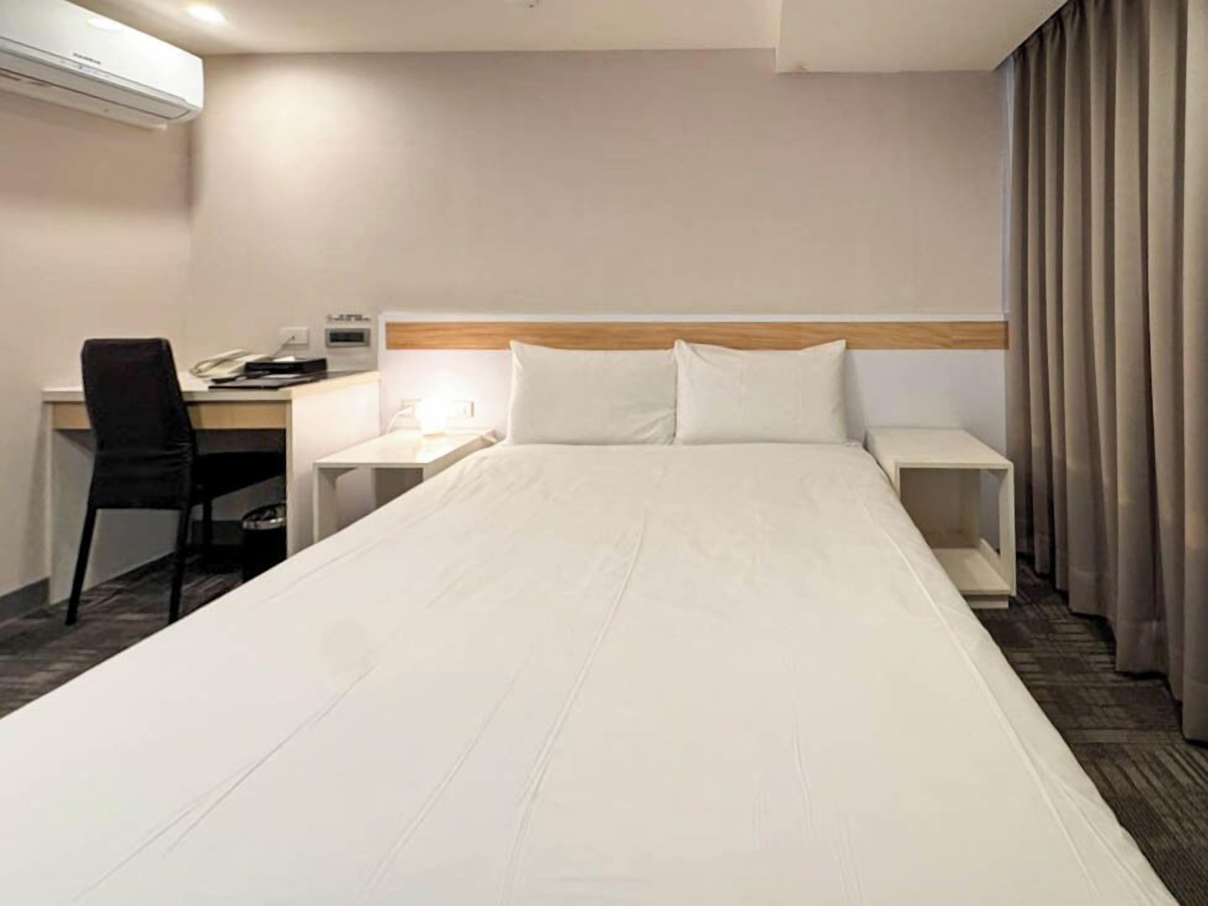 A hotel room with a large bed, a desk, a chair, and a desk lamp.