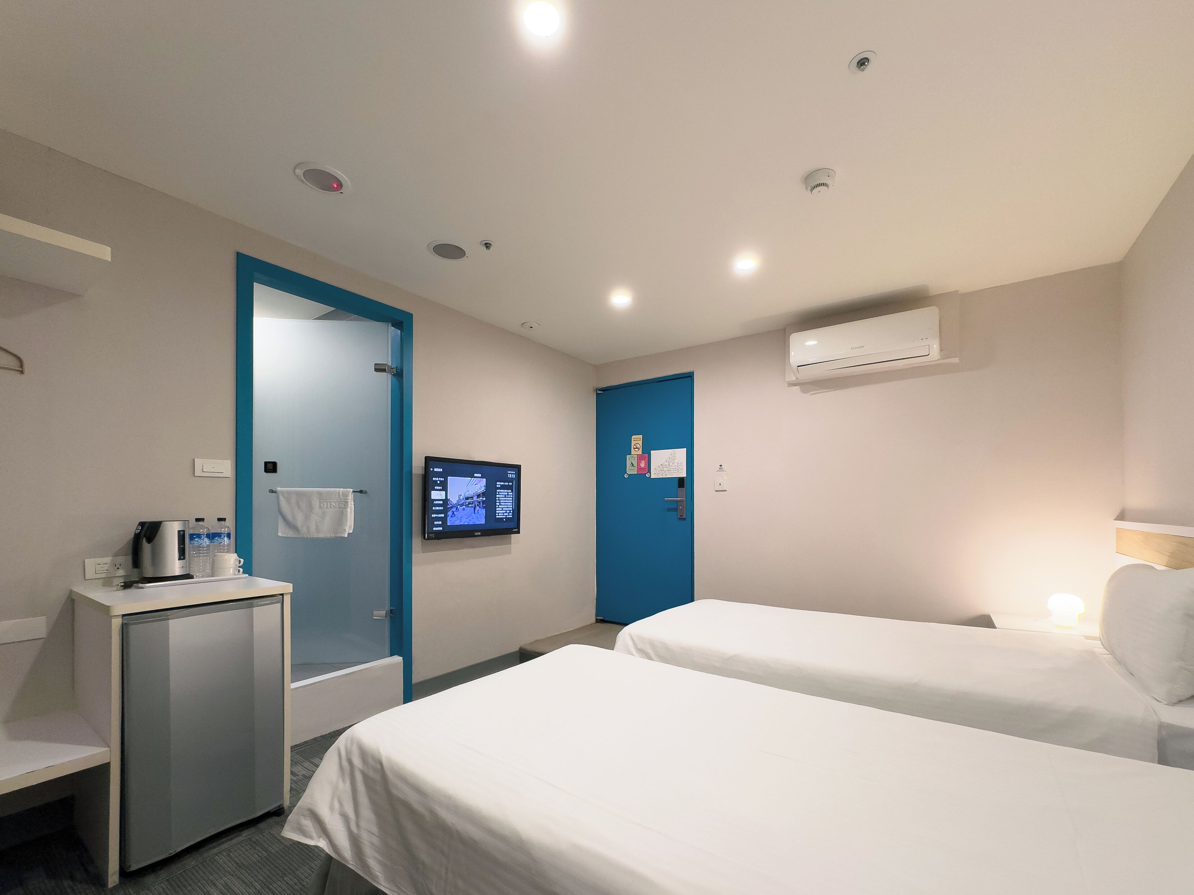 A hotel room with two beds, a desk with a kettle and cups, a TV on the wall, and an air conditioning unit.