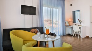 A modern hotel room with a yellow sofa, a small round table with a drink and fruits, a desk with a chair, and a TV mounted on the wall. - Mikes Kanarium Hotel (Larnaca)
