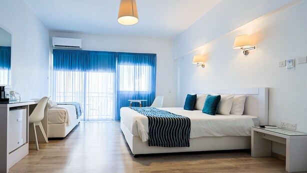 A modern hotel room with a large bed, a desk, and a chair. There is a window with blue curtains and an air conditioning unit on the wall. - Mikes Kanarium Hotel (Larnaca)