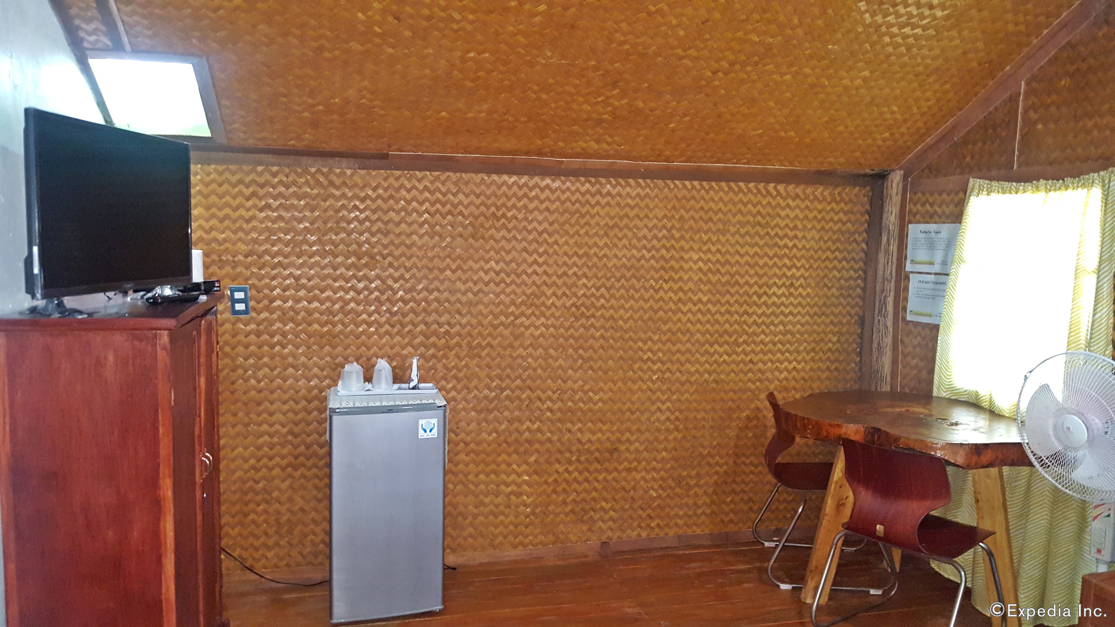 A room with a wooden ceiling, a small refrigerator, a wooden desk with a chair, a television on a cabinet, and a window with curtains.