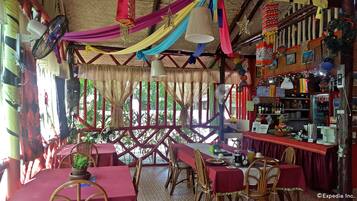Um restaurante com interior rústico, apresentando um telhado de palha, paredes listradas em vermelho e branco e uma variedade de decorações penduradas. Mesas dispostas com cadeiras de vime e toalhas de mesa vermelhas, e o cardápio exibido na parede.