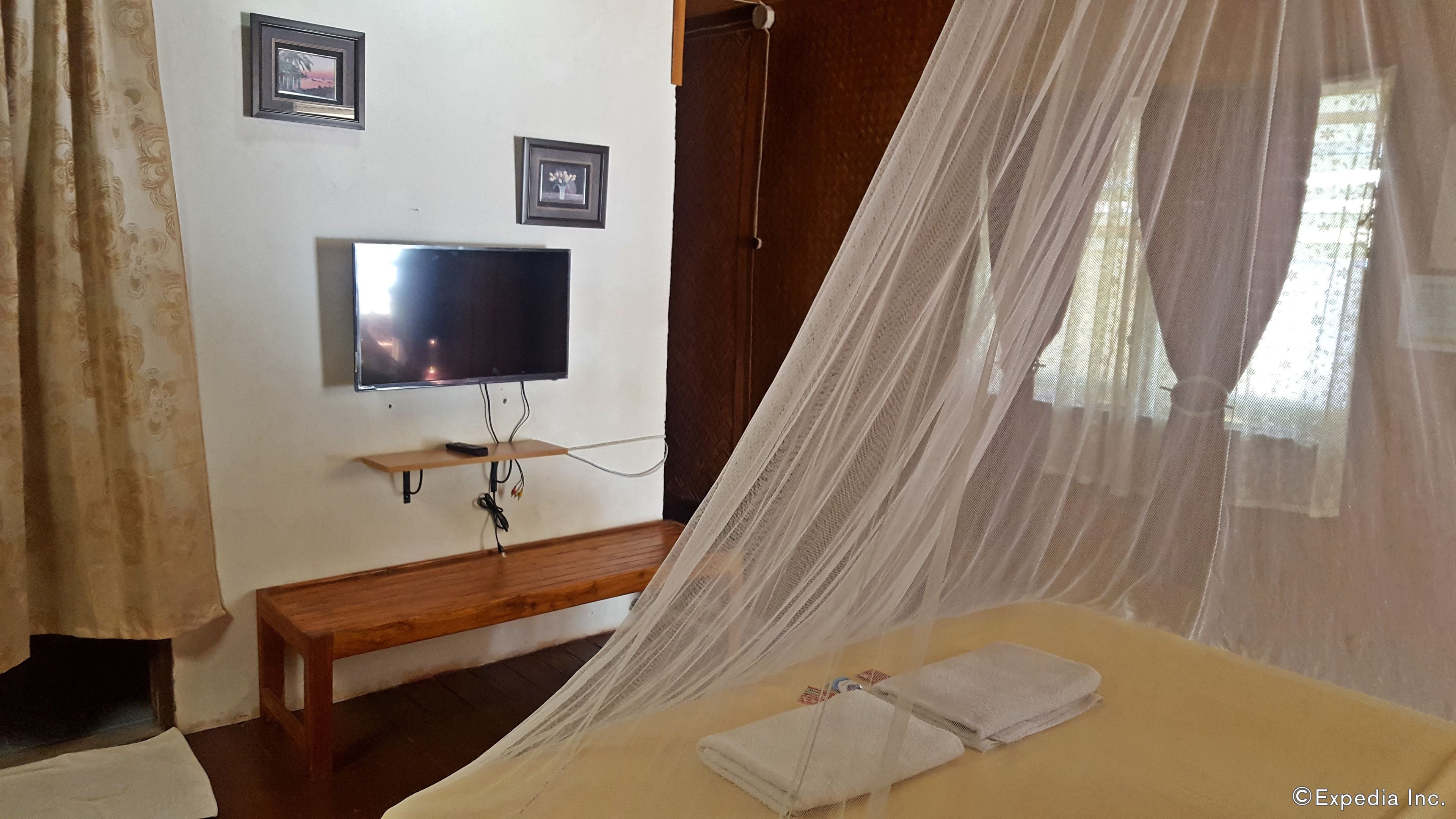 Quarto com cama, televisão na parede, mesa de cabeceira e janela com cortinas leves.