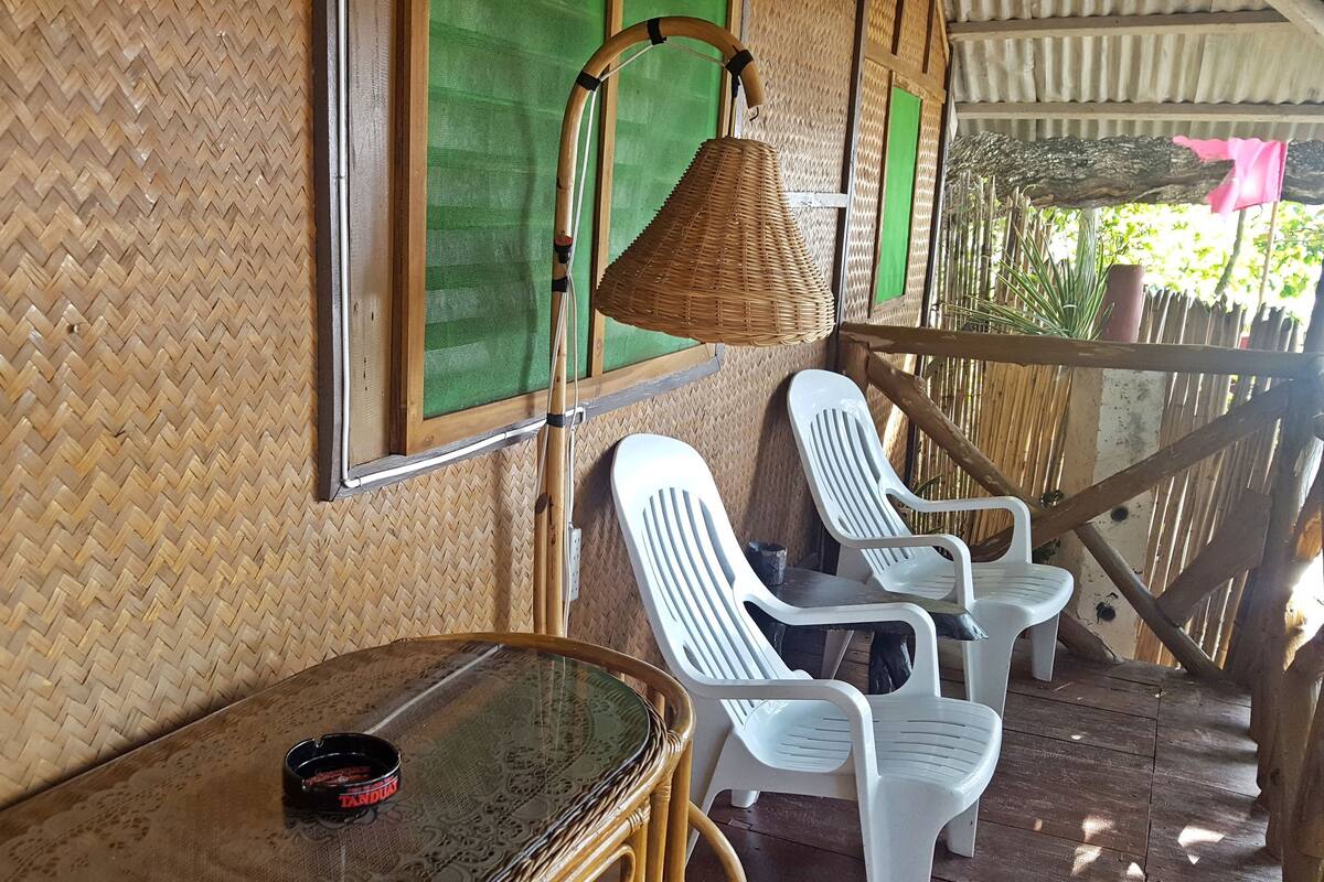 A covered outdoor seating area with woven walls, plastic chairs, a wicker lamp, and a table with a cigarette pack.