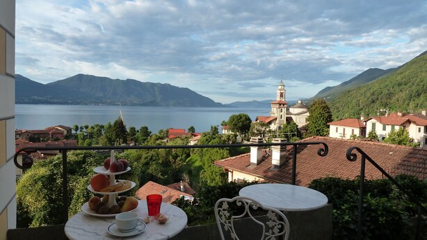 A balcony with a table set for a meal, overlooking a lake and a village with a church tower. - B&B Casa Forster (Cannero Riviera)