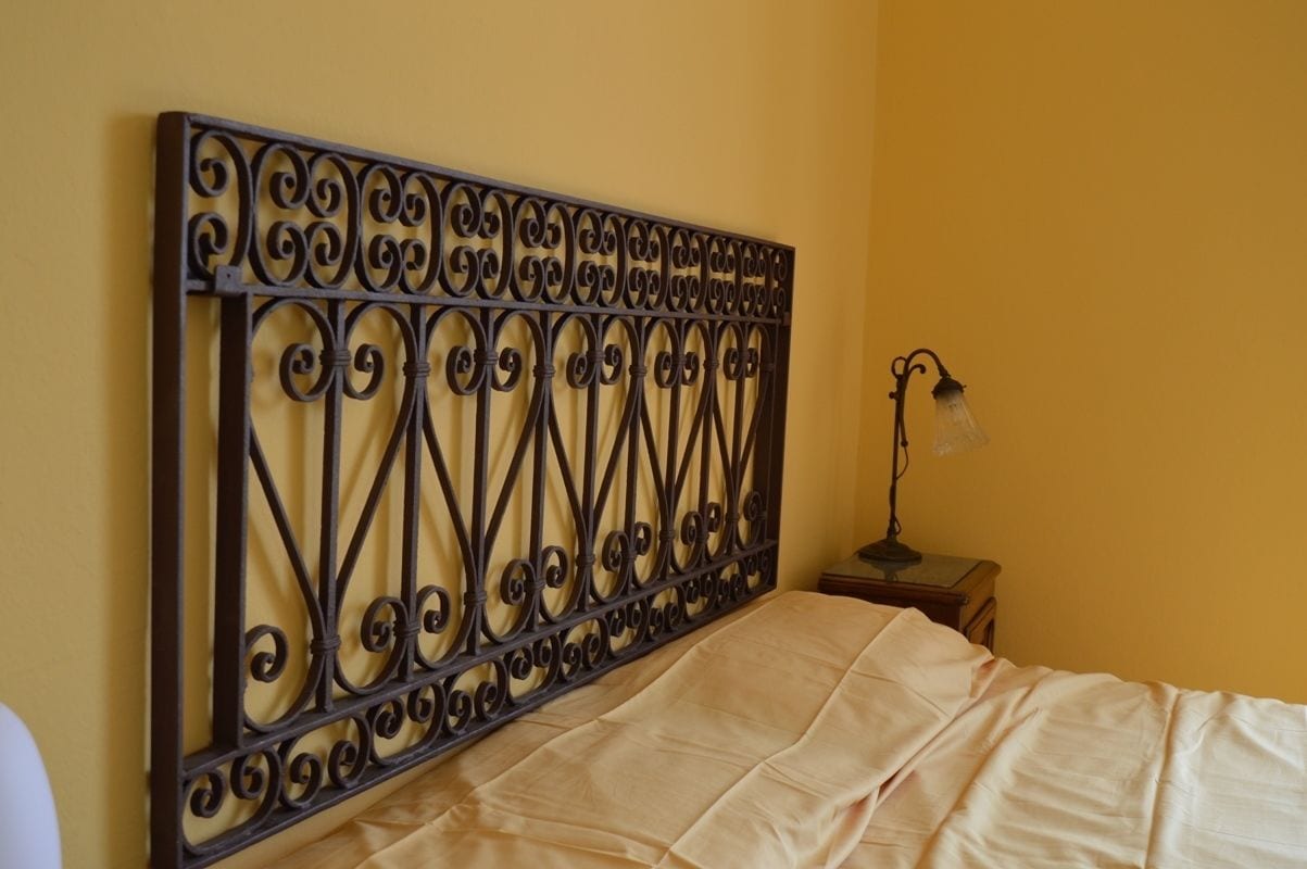 A bed with a headboard featuring intricate metalwork, a bedside lamp, and a plain yellow wall.