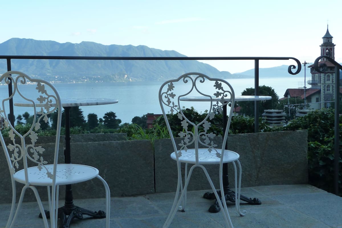 Two ornate white chairs with a round table on a balcony overlooking a body of water and mountains.