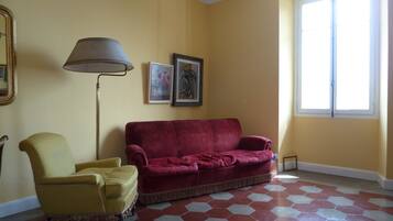 Una habitación con una pared amarilla, un sofá de terciopelo rojo, un sillón amarillo, una lámpara de pie y dos cuadros enmarcados.