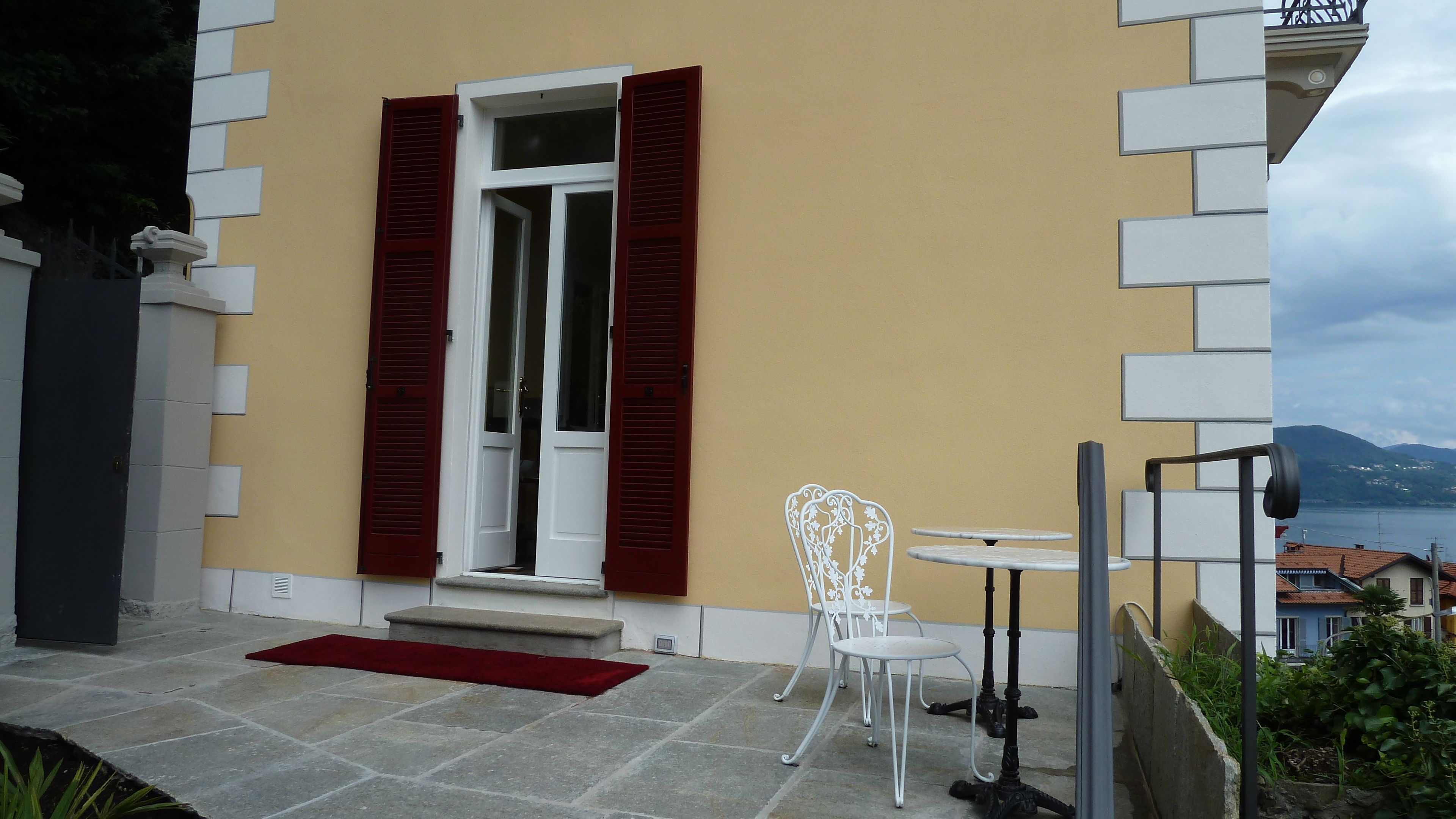 Un edificio moderno con facciata gialla, una porta bianca con persiane rosse e un piccolo spazio esterno con sedie e un tavolo bianchi.