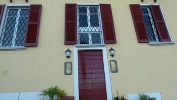 Fachada de edificio con tres ventanas, cada una con contraventanas rojas y rejas metálicas. Hay una puerta grande de rojo oscuro con una pequeña placa encima. Dos faroles de pared se ven a cada lado de la puerta.
