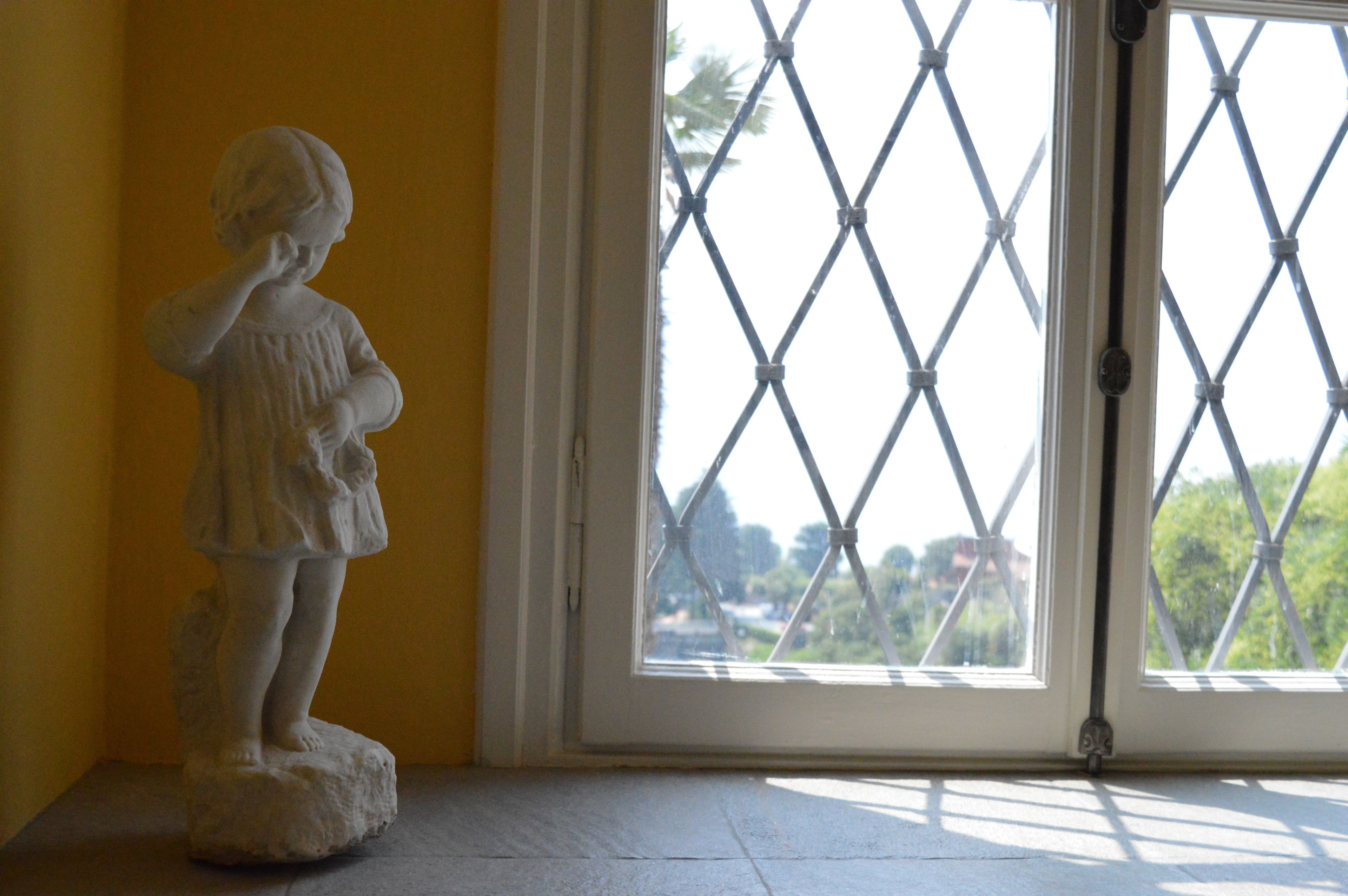 Une statue d’un enfant se trouve dans une pièce dotée d’une fenêtre ornée d’une grille à motifs losangés.