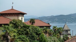 A coastal building with a red-tiled roof, surrounded by lush greenery and overlooking a body of water with distant hills.