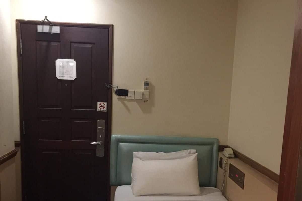 Habitación de hotel con una cama individual, una puerta de madera, un teléfono en la pared y una mesita de noche pequeña.