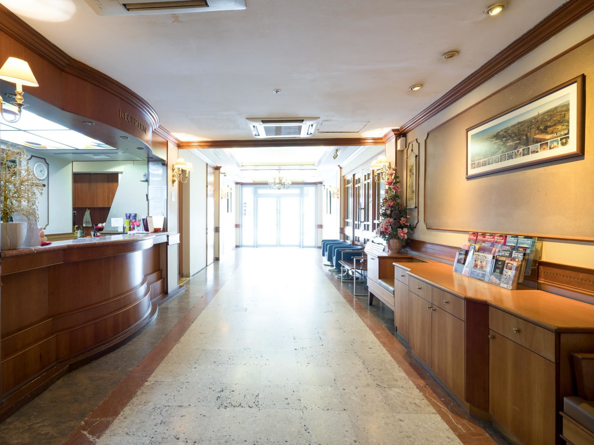 A hotel reception area with a wooden reception desk, a clock, and a framed cityscape picture on the wall.