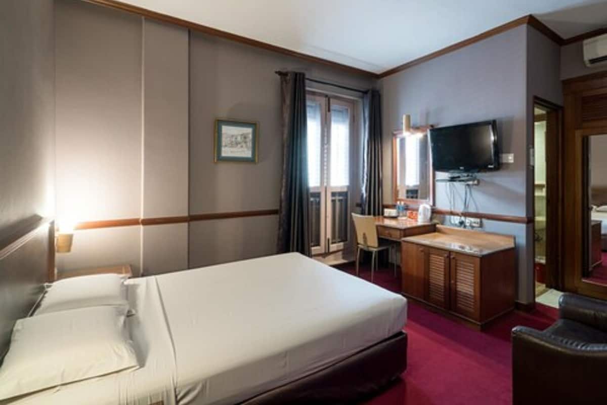 Habitación de hotel con cama, escritorio, silla, televisor y ventana con cortinas.