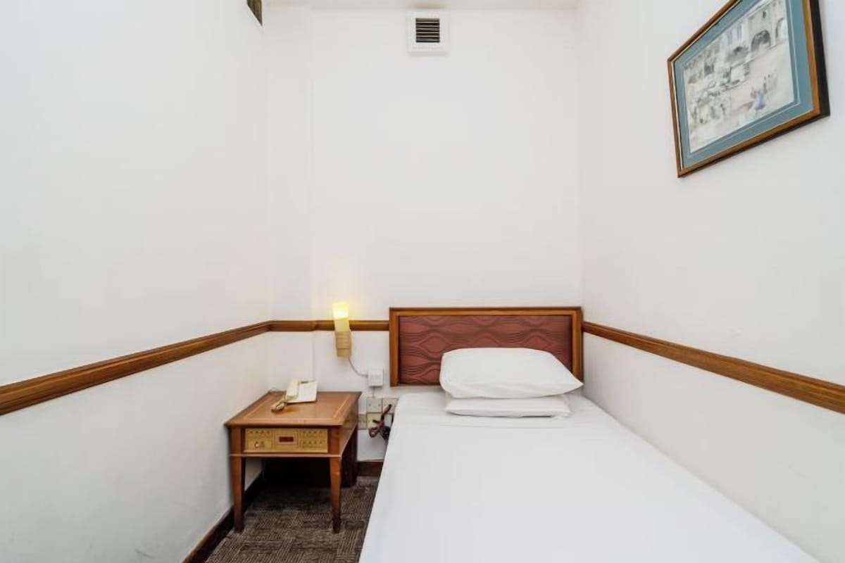 A small hotel room with a single bed, a bedside table with a lamp, a small wooden nightstand, and a framed picture on the wall.